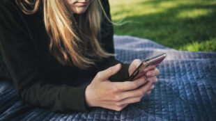 Kvinna ligger på en filt på en gräsmatta och skriver på sin mobil.