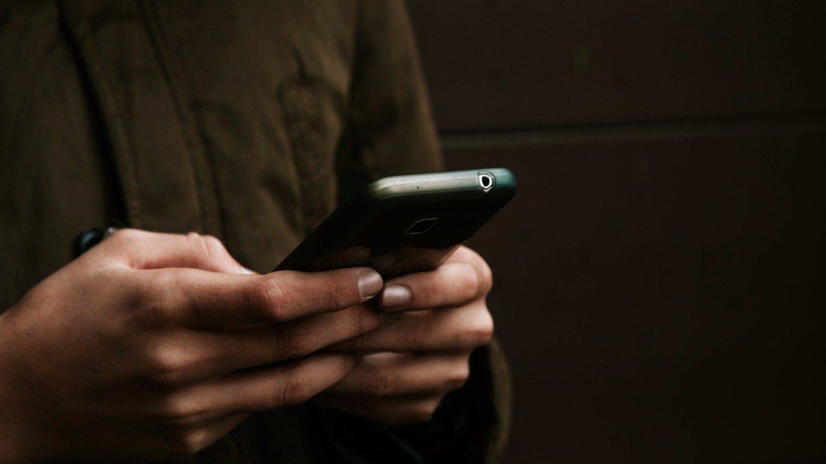 Två händer håller i mobiltelefon, brun skjorta och brun bakgrund