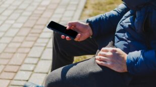 Person med blå jacka och grå byxor sitter utomhus med telefon i handen