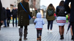 Kvinna och barn går på stenlagd stadsgata, bland andra människor, bara ryggen syns