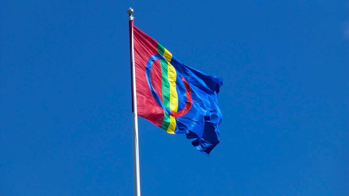 Den samiska flaggan med sina karakäristiska färger (rött, blått, grönt och gult) och mönster, vajar i vinden på en flaggstång.