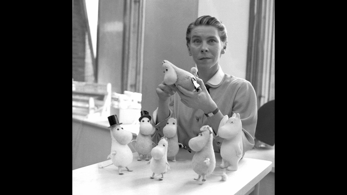 Svartvit bild på Tove Jansson som sitter med Mumintroll-miniatyr i handen. Flera miniatyrer står framför henne på ett bord.