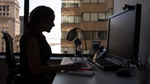 Kvinna, i svart profil, sitter och jobbar vid dator med fönster och stadsmiljö utanför