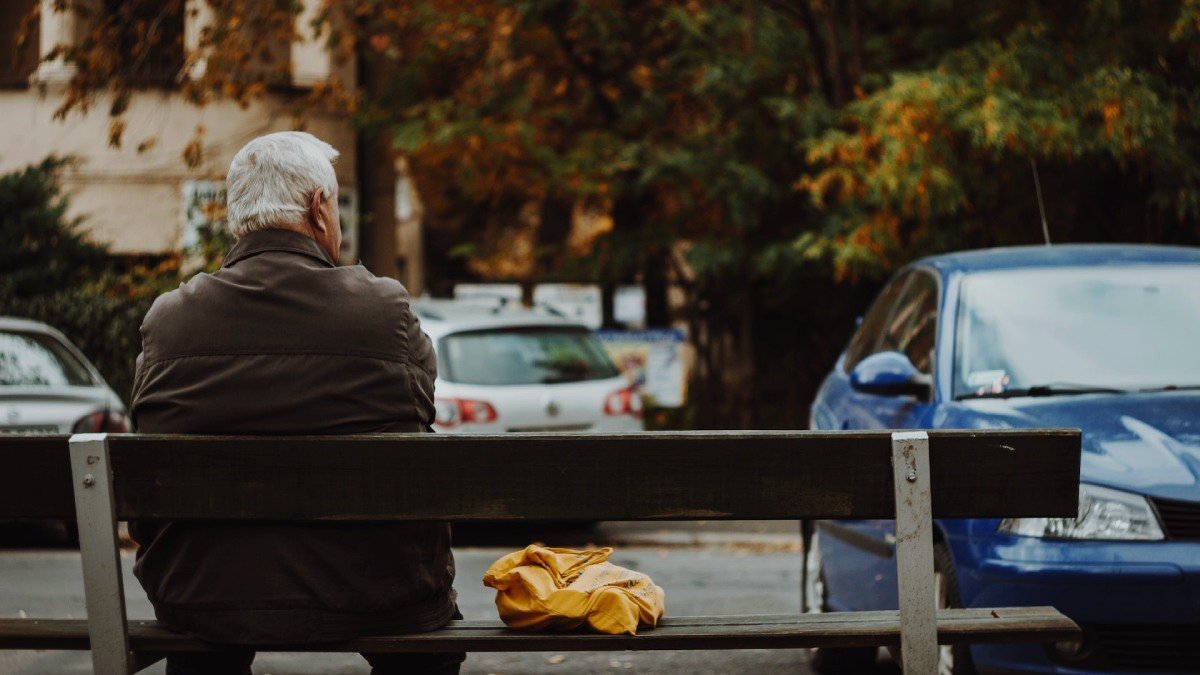 Person med kort grått hår och brun jacka sitter på bänk i stadsmiljö, rygg och bakhuvud syns.