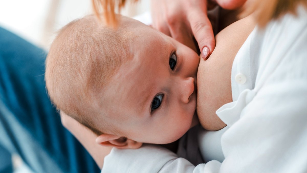 Liten bebis som ammar och tittar in i kameran, mamma i vit skjorta skymtas.