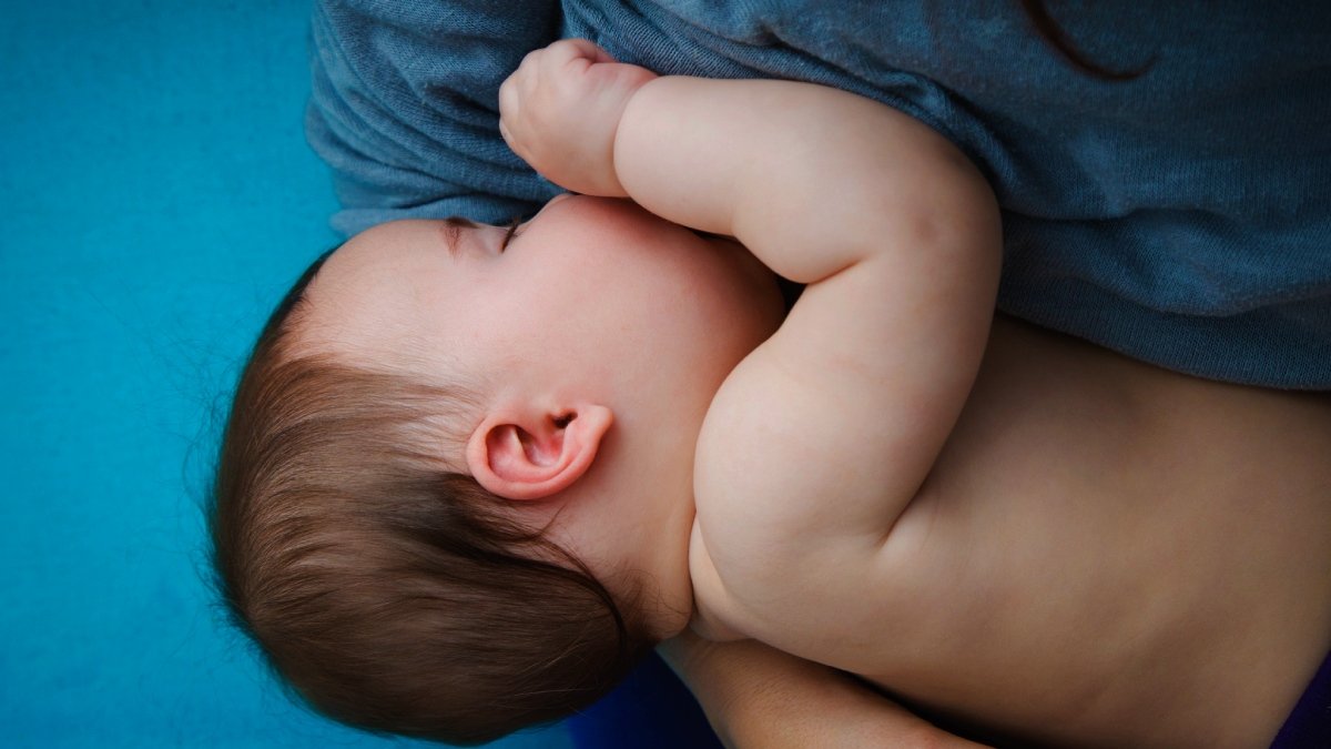 En bebis med bar överkropp som blir ammad, sedd uppifrån, blå skjorta på vuxenkroppen och blå bakgrund.