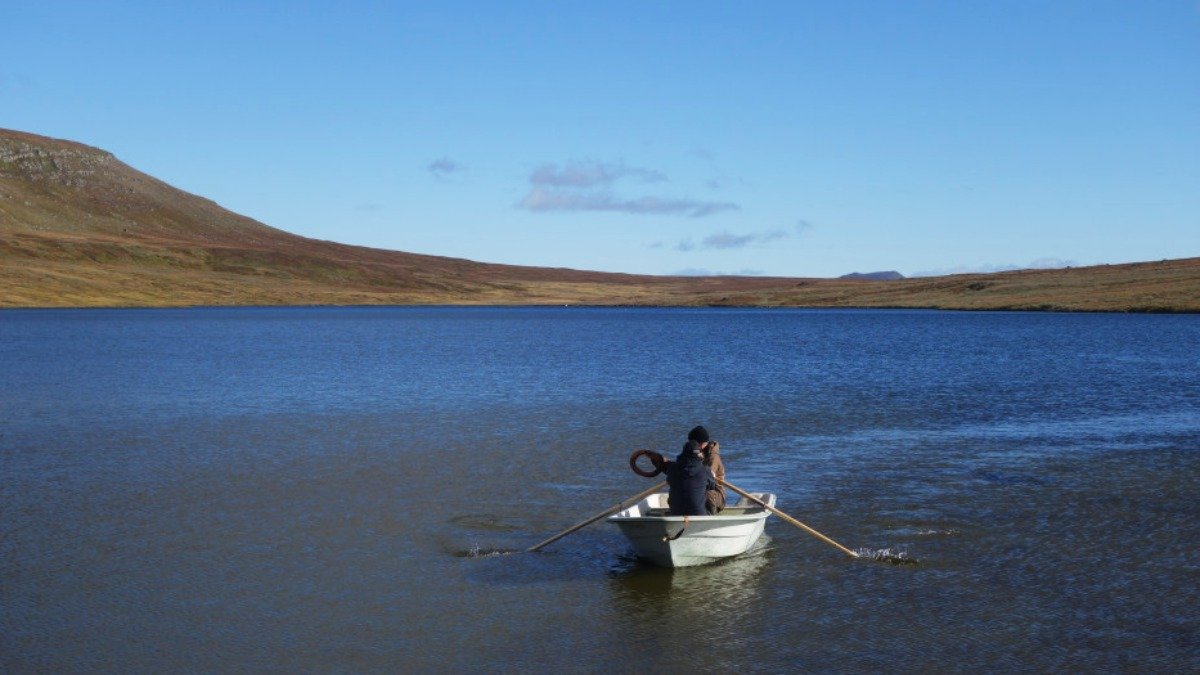 Vit roddbåt med två personer i, på sjö. Berg i bakgrunden,