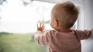 Litet barn står med hand mot fönster, tittar ut, ljust hår och randig tröja.
