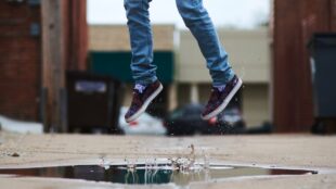 Två barnsben, i jeans och sneaker, i ett hopp ovanför en vattenpöl utomhus.