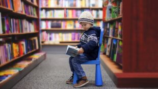 Barn i sexårsåldern sitter på stol omgiven av bokhyllor och tittar i en bok.