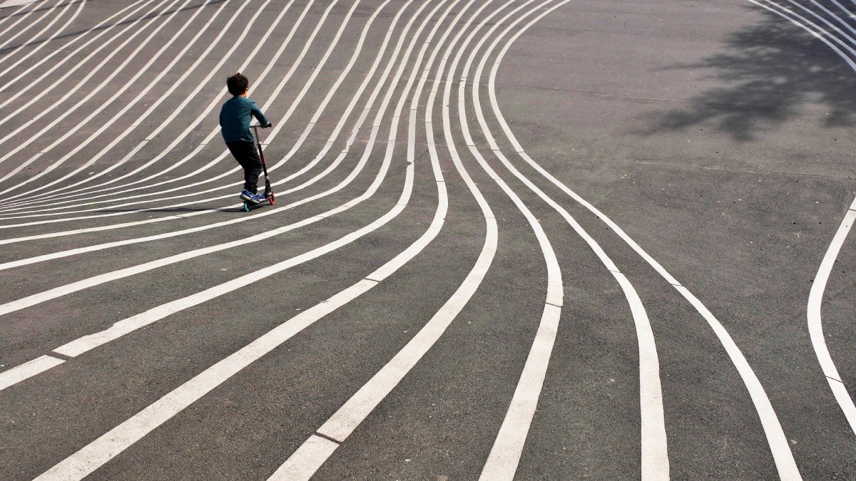 Ett barn åker sparkcykel nedför backe där långa vita streck har målats på den asfalterade marken, som spår att åka i.