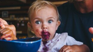 Bebis framför blå matskål, har blått kladd runt munnen, vuxen sitter tätt bakom.