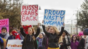 Två unga kvinnor håller upp skyltar med texten "Pussy grabs back" och "Proud nasty woman" under demonstration