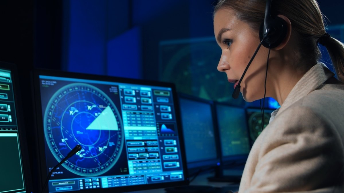 Kvinna med headset, i profil i flygledarmiljö med datorer och skärmar.
