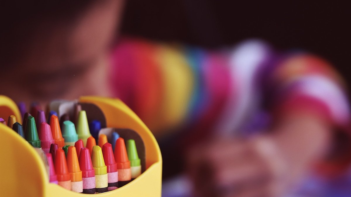 En ask med färgkritor i förgrunden, ett ritande barn i bakgrunden, i oskärpa.