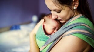 Nyfödd bebis är grönrandig bärsjal på mörkhårig kvinnas bröst.