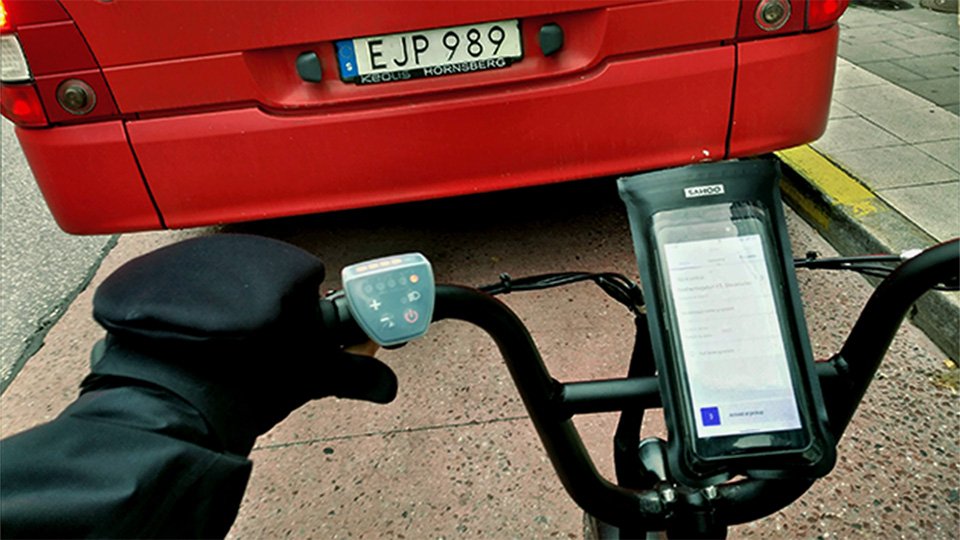 Handskbeklädda händer och mobil med app synlig på skärmen, på ett mopedstyre. Mopeden ligger strax efter en bil som sprutar ut avgaser.