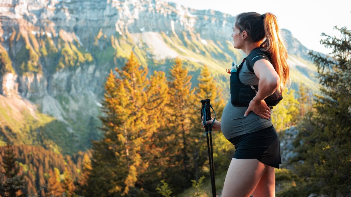 Person med gravidmage står i träningskläder och med gångstav och tittar till vänster. Högt berg och träd med gula löv i bakgrunden.