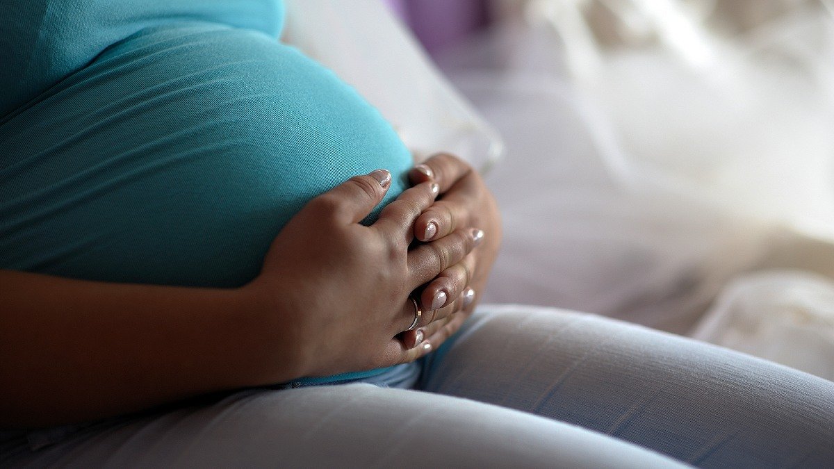 Sittande person håller knäppta händer över sin gravida mage, bär blå kortärmad tröja och grå byxor.