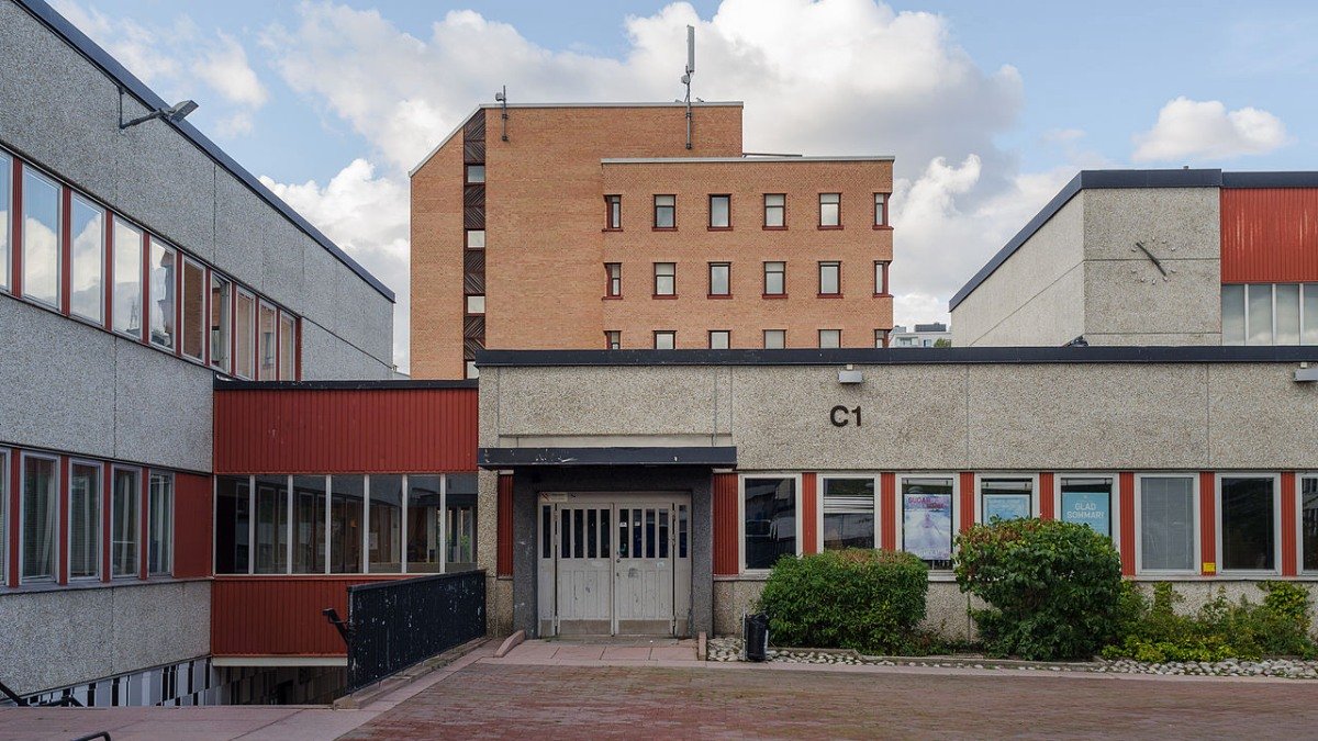 Entrén vid en gymnasieskola i grått och rött. Rött höghus i bakgrunden.