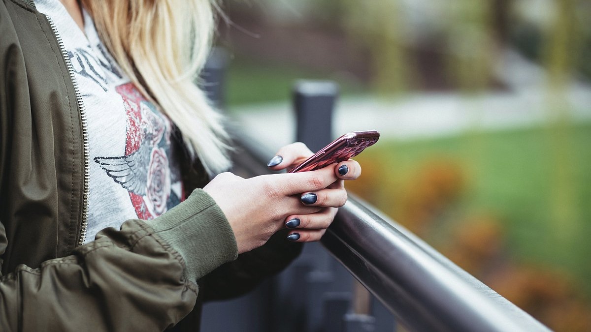 Person med målade naglar håller i mobiltelefon utomhus, inget ansikte, grönska bakom.