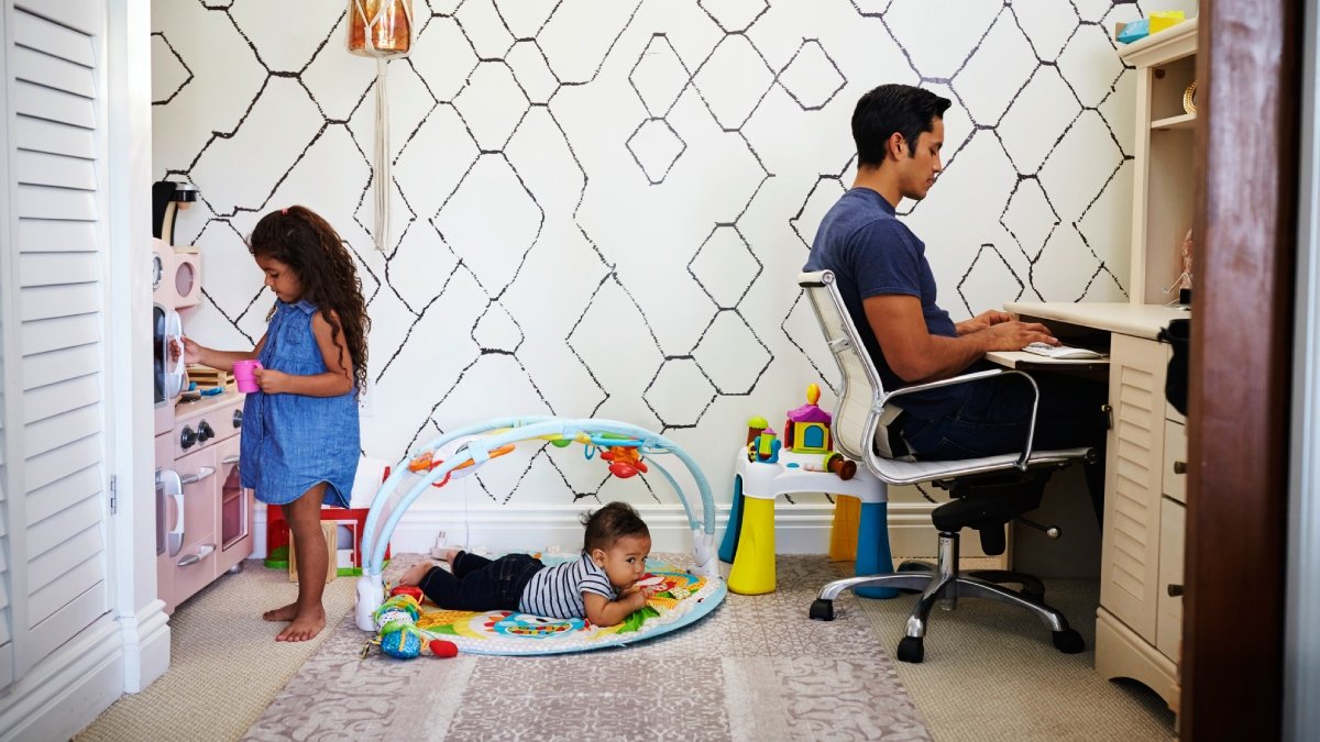 Hemmiljö med liten flicka till vänster, bebis i babygym på golvet i mitten och hemmajobbande pappa till höger, vänd från barnen, vid skrivbord.