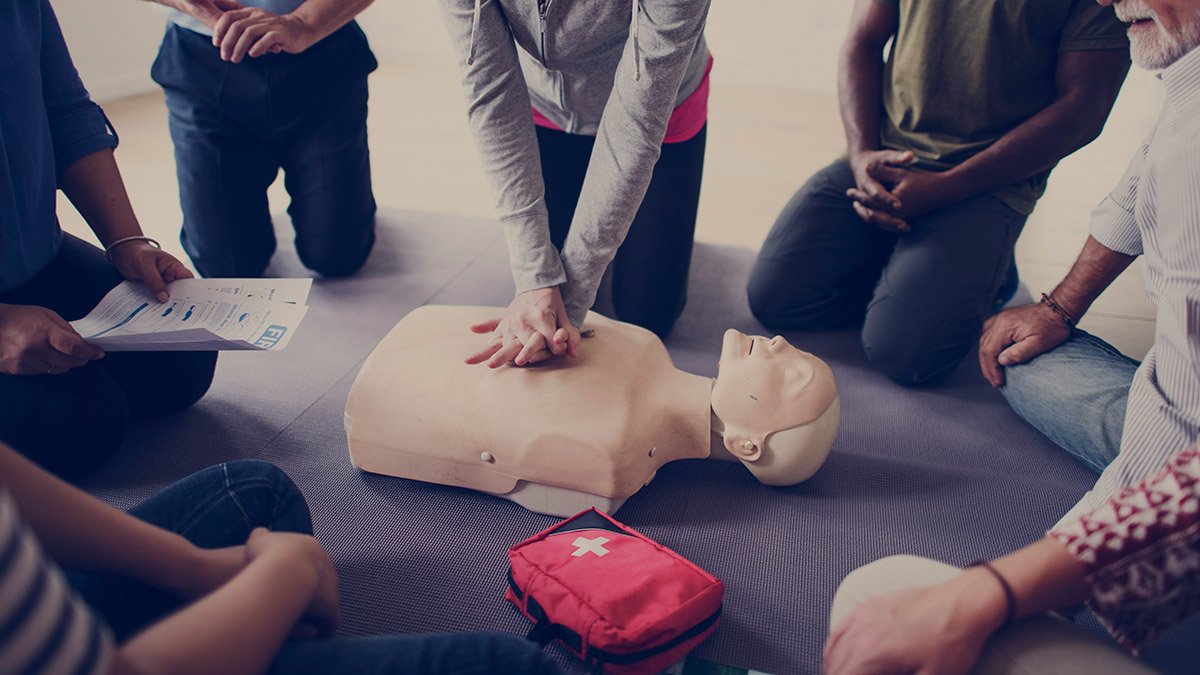 Personer sitter på golvet i en ring runt en testdocka som de tränar hjärt-lungräddning på.