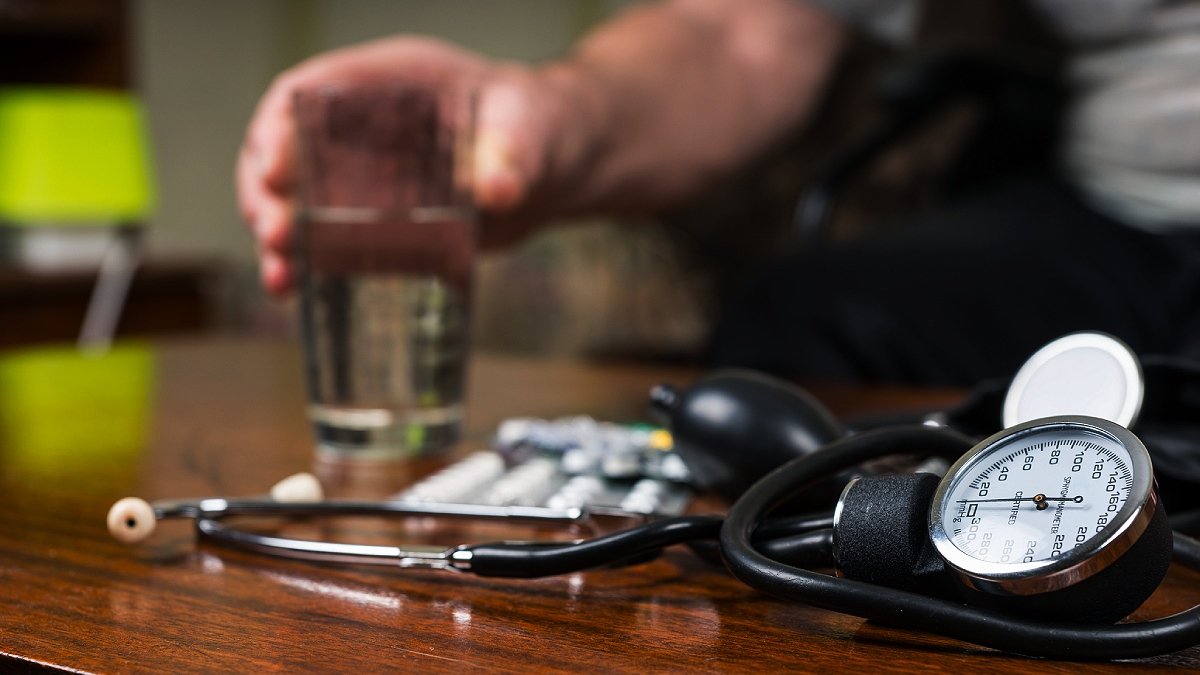 Blodtrycksmätare och piller liggande på träbord, hand med vattenglas i bakgrunden. Om läkmedel vid högt blodtryck.