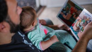 Man med skägg, i profil, läser bok för liten bebis som sitter i mannens famn