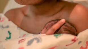Huvud på nyfödd bebis syns delvis bakom täcke, bebisen ligger hud mot hud med en vuxen.