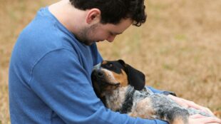 Leende man med mörkt hår och blå tröja kramar hund, utomhus. Hunden och mannen tittar på varandra.