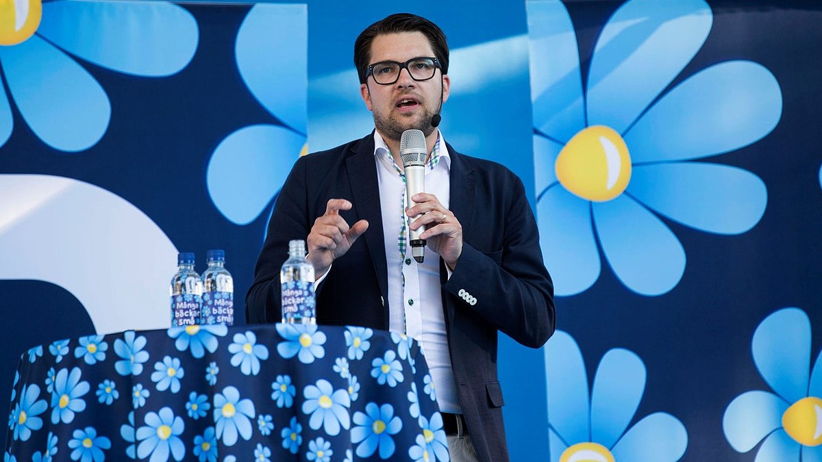 Jimme Åkesson talar, med mängder av SD-blommor på bordsduk och skärm bakom.