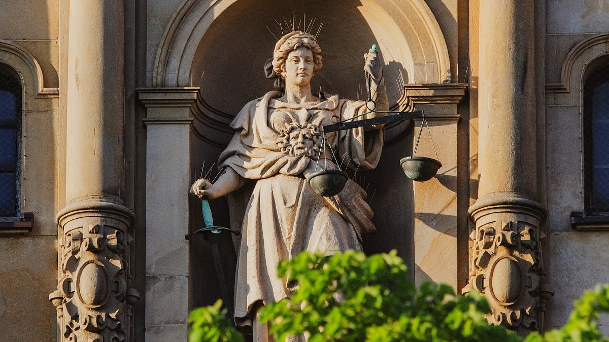 En stenstaty av en kvinna som håller en vågskål i handen, Justitia. Grönska i förgrunden.