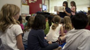 Barn sitter på golvet i klassrum, lyssnar på vuxen som läser bok, ryggar mot kameran.