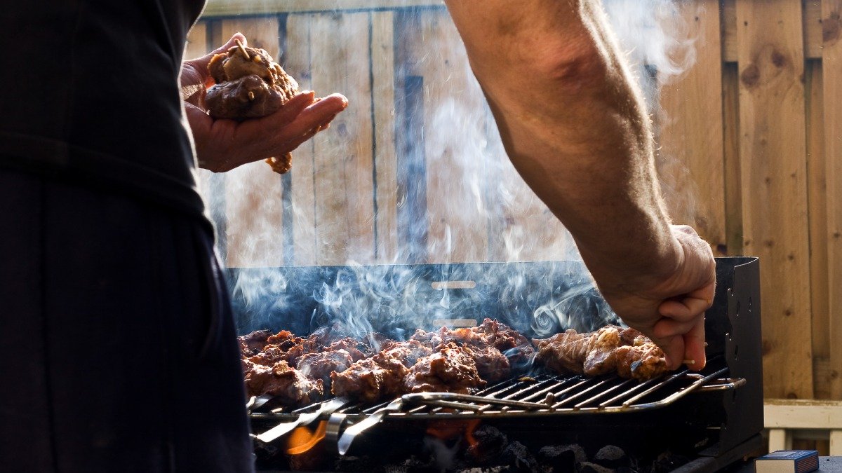 Grill med köttbitar och matos, två armar och lite av en kropp syns invid grillen.