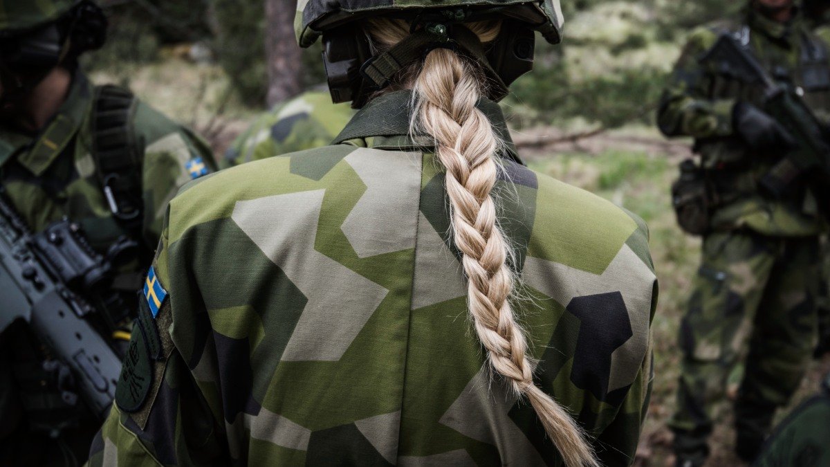 Person med ryggen mot kameran, i terräng och iklädd kamouflagekläder ohc hjälm. Ljus hårfläta tittar fram under hjälmen.