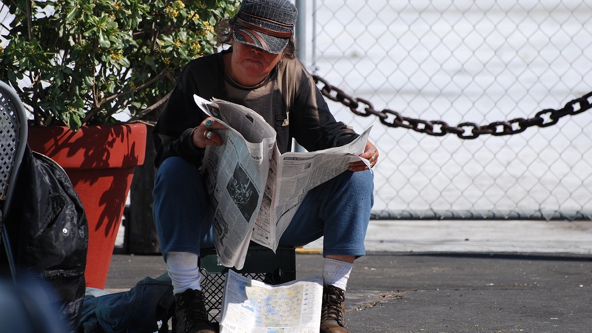 Person sitter utomhus, i keps, och läser dagstidning.