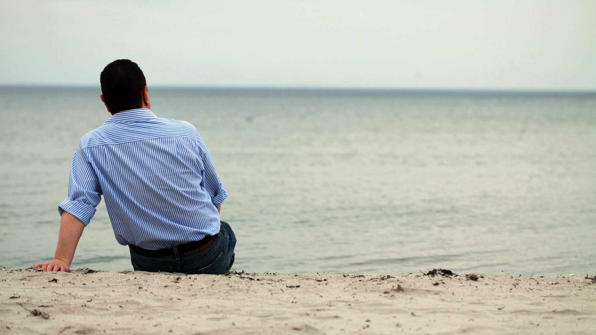 Person i ljus skjorta och med mörkt kort hår sitter med ryggen mot kameran på en sandstrand, vänd mot vattnet.