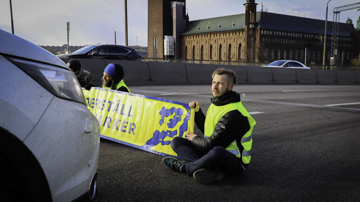 Två personer sitter mitt på en motorväg med en skylt mellan sig.