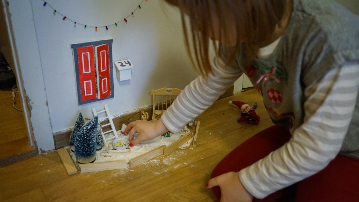 Ett barn sitter på golvet och pysslar med en nissedörr på väggen invid golvsockeln. En minigulgran och låtsassnö finns på golvet.