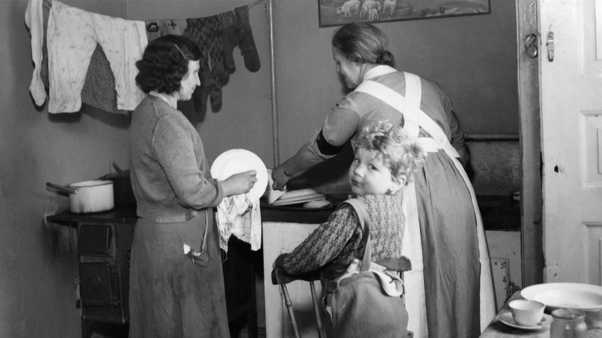 Svartvitt fotografi från 1950-talet. Två kvinnor torkar disk i ett fattigt hem med vedspis. I förgrunden syns en liten pojke som tittar mot kameran.