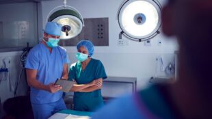 Digitalisering i vården och risker: Två personer i munskydd och blå kläder står och tittar på en digital platta i en operationssal.