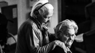 En äldre kvinna står bakom en kvinna som sitter ner och har händerna på hennes axlar.