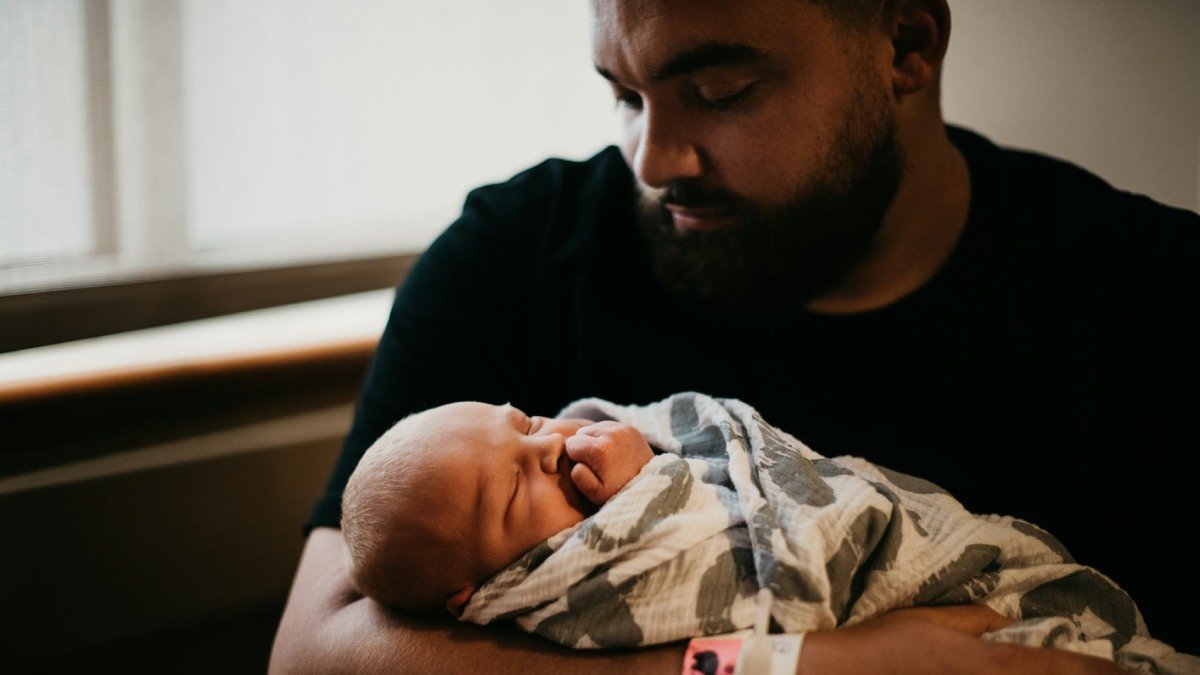 Man med skägg håller nyfödd bebis som är inlindad i filt, sjukhusmiljö.