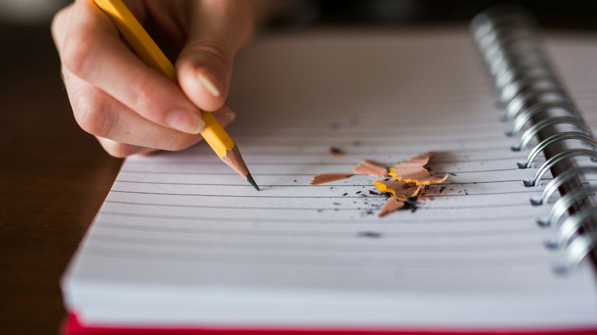 Närbild på hand som håller i blyertspenna som är riktad mot ett anteckningsblock. Rester från pennvässning ligger på pappret.