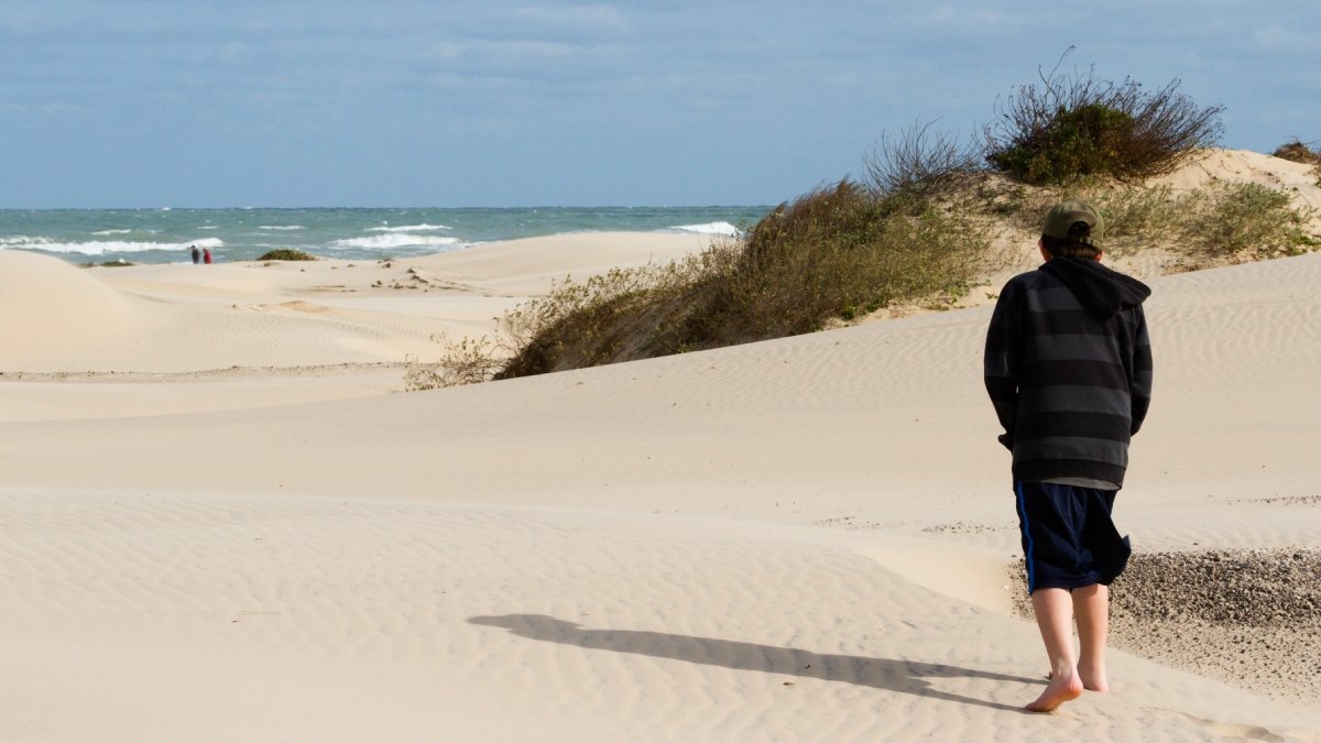 Ung person i mörka kläder går barfota på sandstrand.