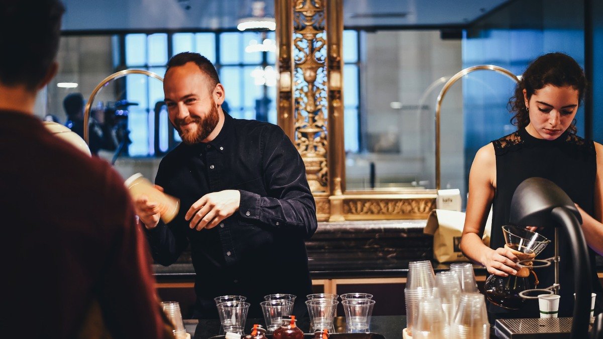 Två personer står bakom en bardisk och blandar drinkar, restaurangmiljö. Personen till vänster ler.