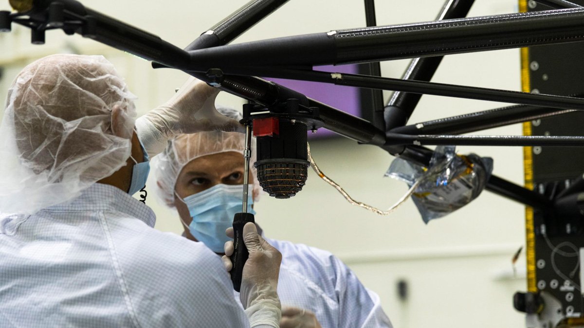 Två personer med skyddande hättor, munskydd och plasthandskar använder skruvmejsel för att montera en sensor på rymdfarkost.