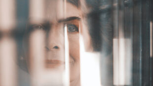 Ansiktet av en äldre kvinna med makeup syns bara delvis genom ett fönster med ljusreflexioner.