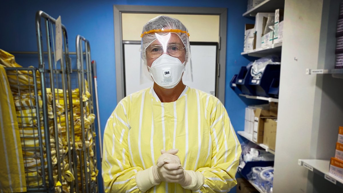 Person står i gulvit skjorta, vita handskar, munskydd, mössa och visir, med händerna knäppta i något slags förråd, sjukhusmiljö.